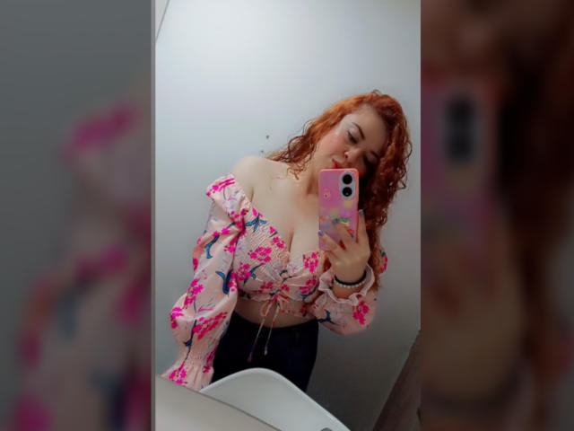 RosseHot6f takes a selfie in a mirror. She has curly red hair and is wearing a pink floral off-shoulder top that reveals her cleavage and black pants. Her pink phone with a floral case is in her right hand. The mirror reflection shows her from the chest up. The composition is centered, with a focus on her upper body and face. The image has a casual, modern style. The colors are bright and vibrant. The texture of her top is slightly loose and floral. The reflection is clear and sharp.