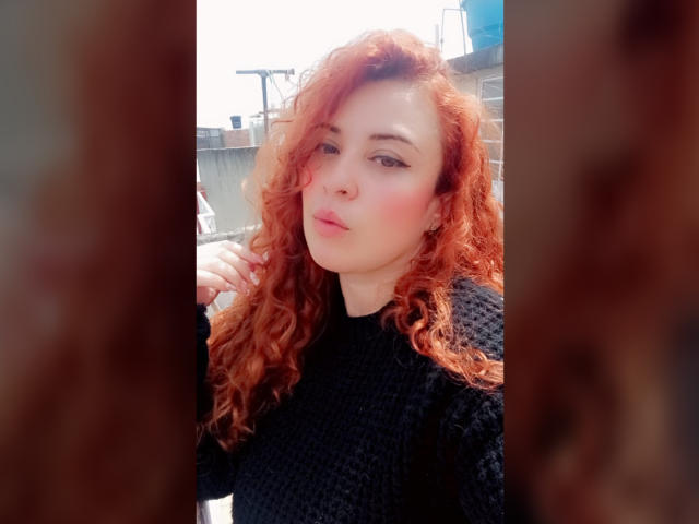 RosseHot6-9 has curly red hair, wearing a black textured sweater. Her skin is fair, with a slight blush on her cheeks. She has a nose piercing and looks directly at the camera with a neutral expression. The image is framed by blurred brown bars on either side. The background is out of focus, showing an urban setting with a blue sky. The lighting is natural, highlighting her hair and face. The composition centers RosseHot6-9, making her the clear focal point.