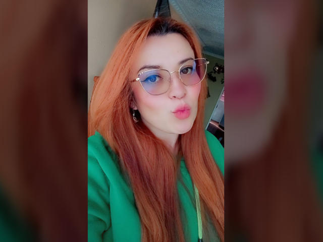 RosseHot69 mit langen roten Haaren, trägt eine grüne Kapuzenjacke und eine Brille, mit leicht gerunzelten, rosafarbenen Lippen, reflektierende Brille, zarten, rosafarbenen Lippenstift, verschwommenes braunes Gesicht im Vordergrund, Innenaufnahme, im Selfie-Stil..