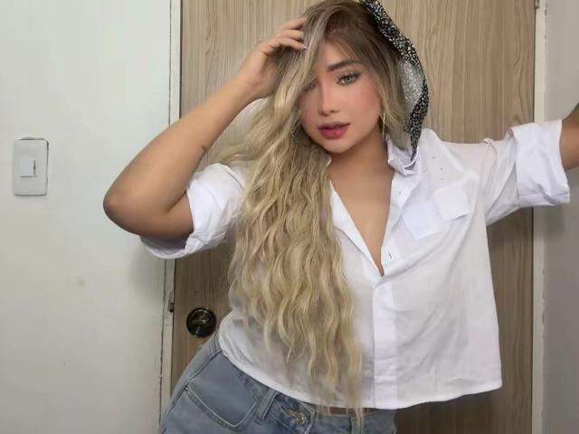 VictoriiaGrey with long blonde hair, white button-up shirt, blue jeans, and black polka-dot hood, touching her head, standing in front of a wooden door.