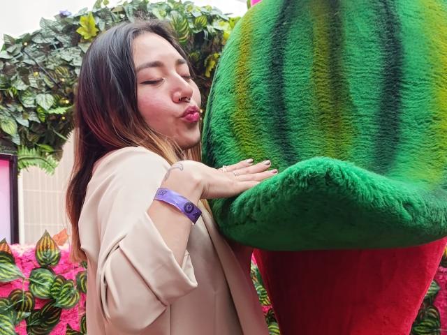 SeleneAna with puffy pink lips, kissing a large green and red fuzzy watermelon costume, wearing a beige top and purple bracelet, in a colorful, plant-filled setting.