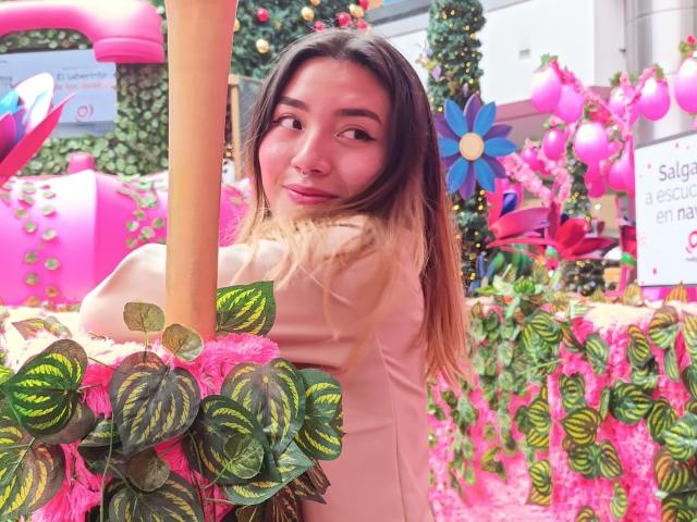 SeleneAna with long brown hair, wearing a pinkish-beige top, stands near a pink column, surrounded by pink flowers, green leafy plants, and colorful decorations, smiling at the camera.