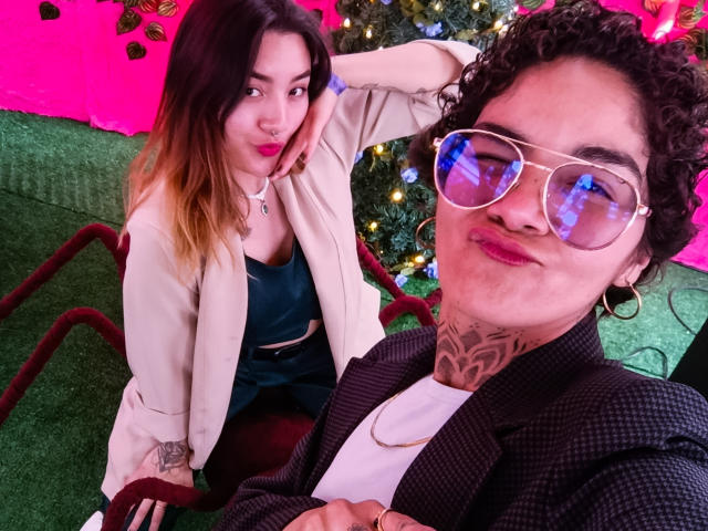 SeleneAna with ombre hair, pink lips, and beige jacket, sitting on red chair. Curly-haired SeleneAna in pinstripe jacket, white tee, and reflective sunglasses, tattoos on neck.