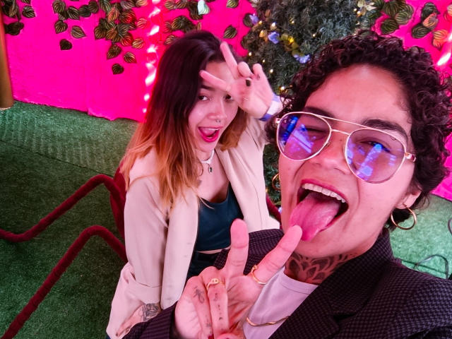 SeleneAna with long, ombre hair, wearing a beige jacket, making a peace sign, and smiling. A curly-haired man with tattoos, sunglasses, and a tongue out, pointing at her.