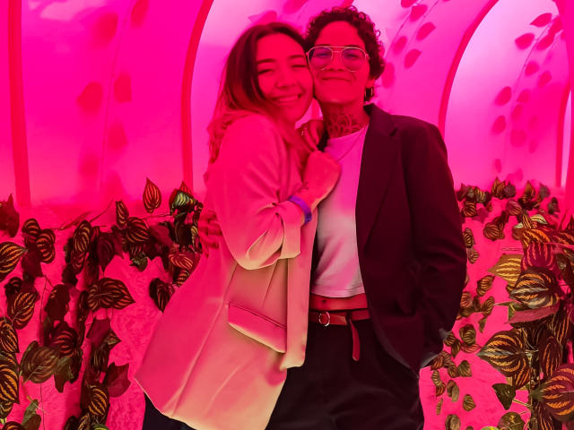SeleneAna with brown hair, pink coat, smiling, arm around tattooed guy in black suit, white shirt, pink neon light. Her tits are small, his cock is not visible.