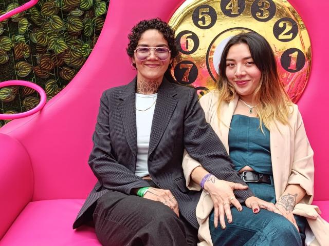SeleneAna with curly hair, glasses, and tattoos in a black pinstripe suit, sitting next to SeleneAna with blonde ombre hair, wearing a blue dress and beige coat.