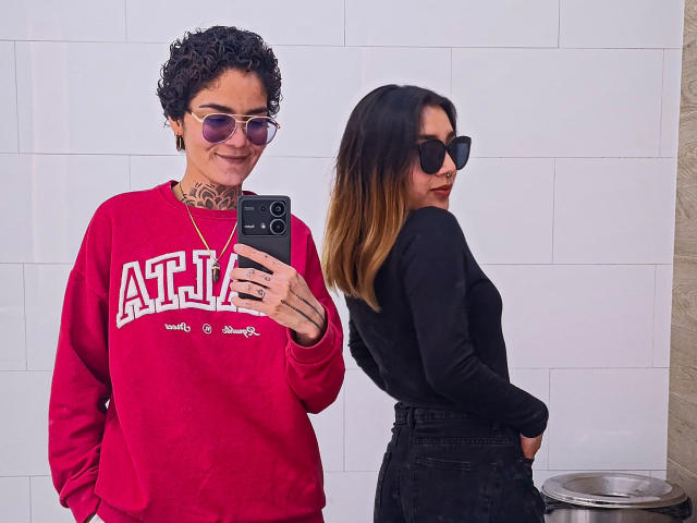 SeleneAna with curly black hair, wearing red sweatshirt and sunglasses, takes a mirror selfie. SeleneAna with black top, brown ombre hair, and black sunglasses, stands to the right.