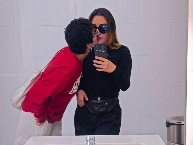 SeleneAna in black top and black pants, taking mirror selfie with phone. Curly-haired dude in red shirt and white pants kisses her cheek. White bathroom, metal trash can.