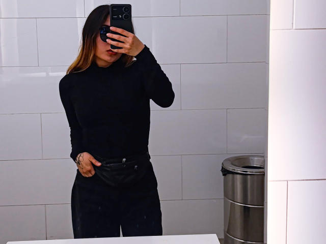 SeleneAna with long brown hair, wearing a black long-sleeve top and black pants, taking a mirror selfie in a white-tiled bathroom, hand in pocket, phone in front.