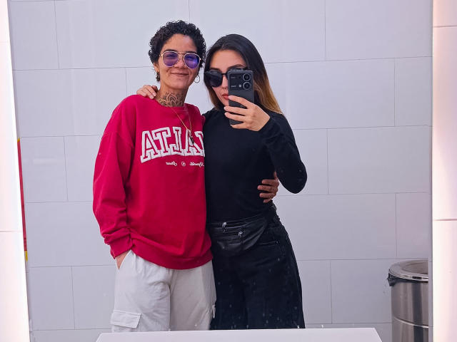 SeleneAna with curly black hair, purple sunglasses, red "ATHLETICS" sweatshirt, white pants, tattooed neck, standing arm around SeleneAna in black top, black skirt, taking mirror selfie.