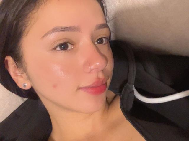 Kloe Thompson ha la pelle color nocciola, i capelli neri, un piccolo piercing al naso, sopracciglia scure e un leggero sorriso con labbra rosate. Indossa una giacca nera con bordi bianchi..
