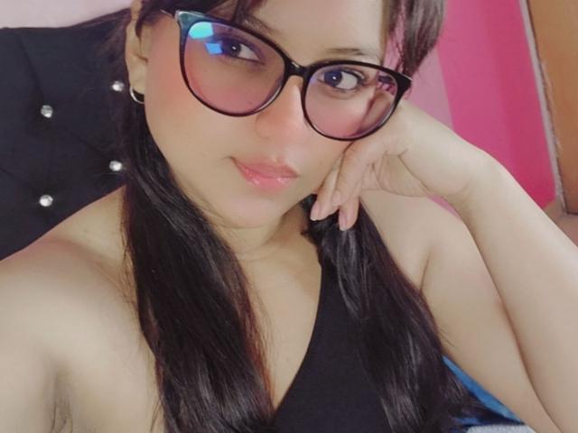 AzulMoon, usando óculos, com cabelo preto e lábios rosa, com a cabeça apoiada na mão, mostrando seus ombros nus e uma blusa preta, olhando levemente para cima. Seus seios estão parcialmente visíveis..