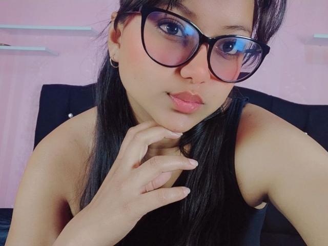 AzulMoon, com cabelo escuro, óculos e lábios rosados, tocando o queixo, vestindo uma blusa preta sem mangas, mostrando sua pele lisa e bronzeada e seu rosto bonito e sensual..