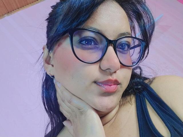 AzulMoon has dark blue eyes, black glasses, and black hair, with pink lips and a hand on her cheek. She's wearing a black top, showing her tits.
