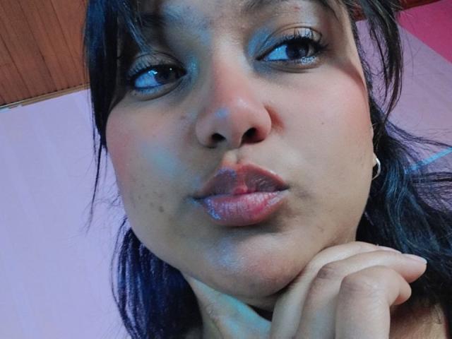 Los labios carnosos y brillantes de AzulMoon están ligeramente entreabiertos, y sus ojos oscuros miran hacia un lado, con una mano apoyada debajo de la barbilla. Su piel está ligeramente brillante, con pequeñas manchas de acné..