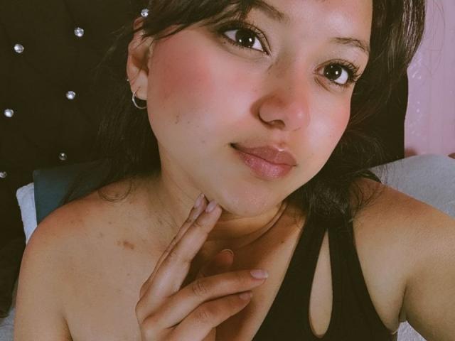 A AzulMoon tem um rosto fofo e rechonchudo, com bochechas rosadas, olhos castanhos e cabelo preto. Ela está vestindo uma regata preta e tocando o queixo com um dedo..