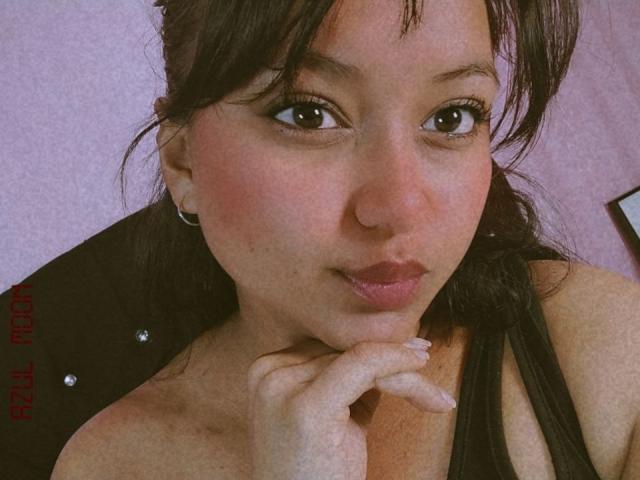 AzulMoon has puffy pink lips, dark brown eyes, and slightly flushed skin. She's biting her finger with black hair and small earrings, wearing a black top.