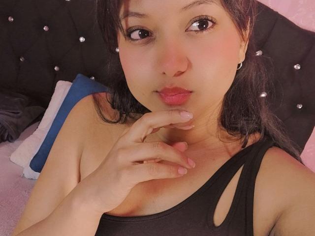 AzulMoon tiene labios rosados y finos, ojos marrones y cabello negro, y lleva un top negro con detalles recortados, tocándose la barbilla con los dedos, sobre una alfombra rosa..