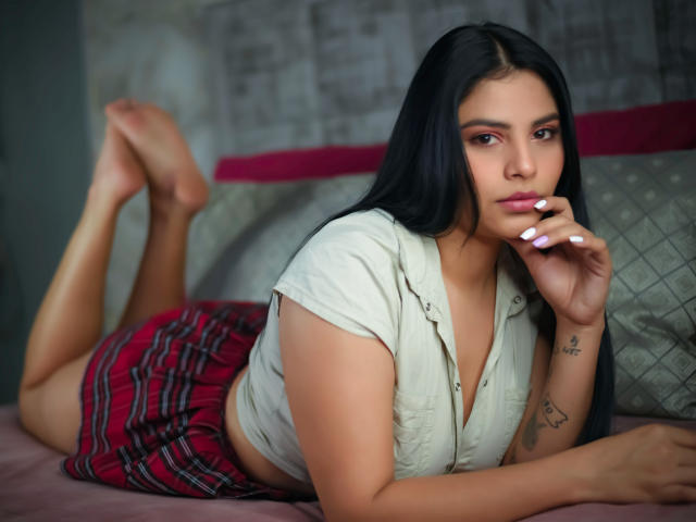 HarmonyBecker with long black hair, laying on bed, white shirt, red plaid skirt, finger on lips, tattoo on right arm, white nails, bare feet, looking at camera.