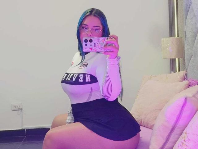 AuroraSantorini with blue hair, glasses, white long-sleeve top, black skirt, sitting, holding phone, showing curvy tits and thick thighs, pink and white pillows in background.