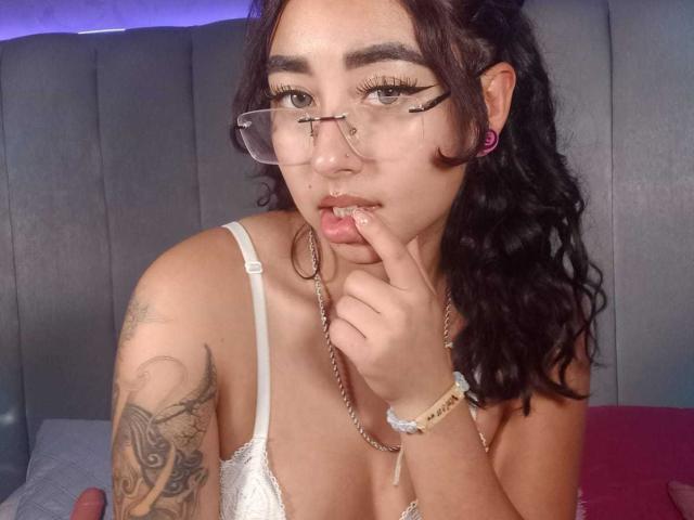 AryVixen with glasses, white lace bra, finger in mouth, brown wavy hair, tattooed arm, pink hoop earrings, chain necklace, and bracelet, in a bedroom.