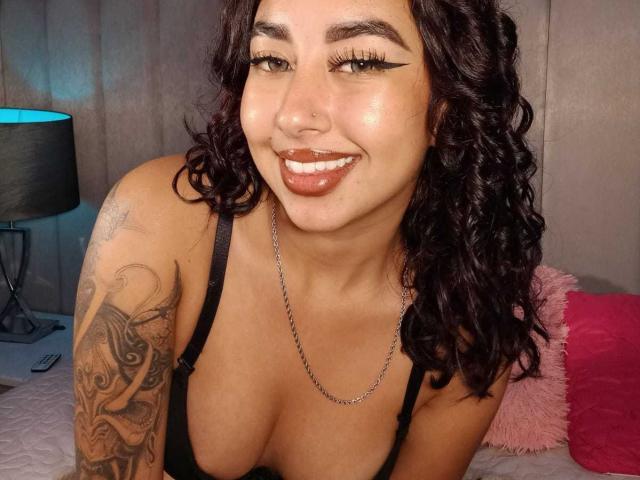 AryVixen met krullend donker haar, een neusring en een getatoeëerde arm, lachend met donkere lipstick, een zwarte beha en een zilveren ketting, waarbij haar borsten zichtbaar zijn, zittend op een bed..