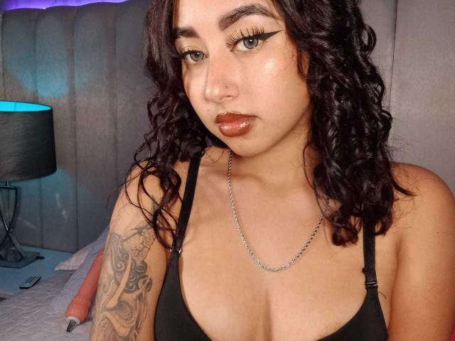 AryVixen, con cabello castaño oscuro y rizado, vestida con una camiseta negra, mostrando su pecho y un tatuaje en su brazo izquierdo, tiene labios brillantes y un collar de cadena..