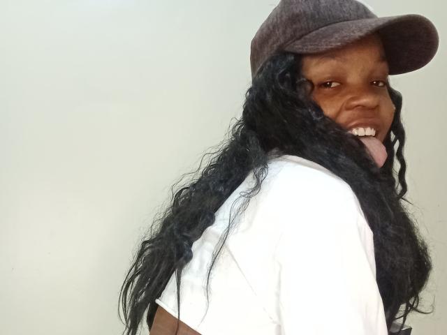 AllaTracy, con piel oscura, cabello negro largo y rizado, camiseta blanca, gorra marrón, sacando la lengua y sonriendo. Su trasero no es visible. Está mirando por encima del hombro..