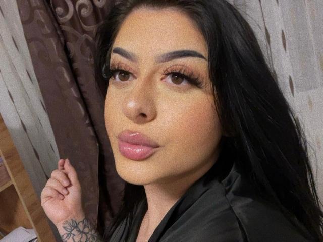 NayaNoir has glossy pink lips, dark eyebrows, long black hair, and is wearing a black shirt, with a tattoo on her right hand, looking at the camera.