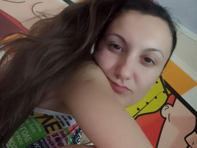ZafiraMoonn's face is close to the camera, with long brown hair over her shoulder, looking slightly annoyed. She has light skin and brown eyes, wearing a colorful top.