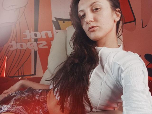 ZafiraMoonn with long brown hair, white ribbed crop top, and plaid pants, showing her flat stomach and small tits, sitting in a room with warm red lighting.
