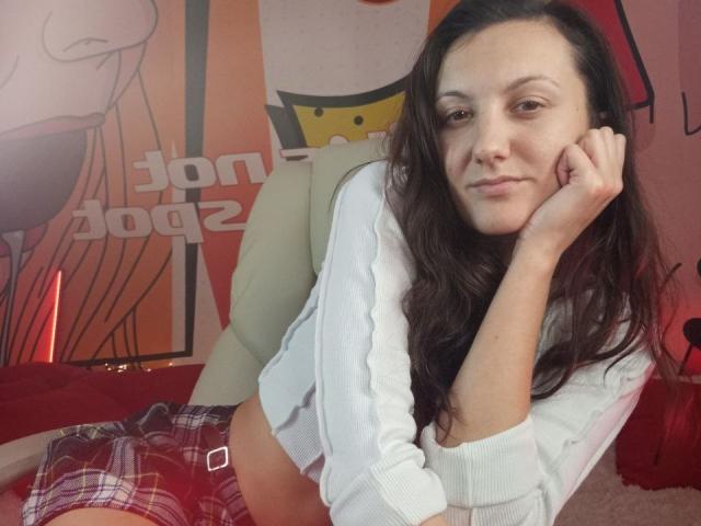 ZafiraMoonn with long brown hair, white ribbed shirt, and plaid pants, resting her chin on her hand, looking at the camera, showing a subtle smile, in a relaxed pose.