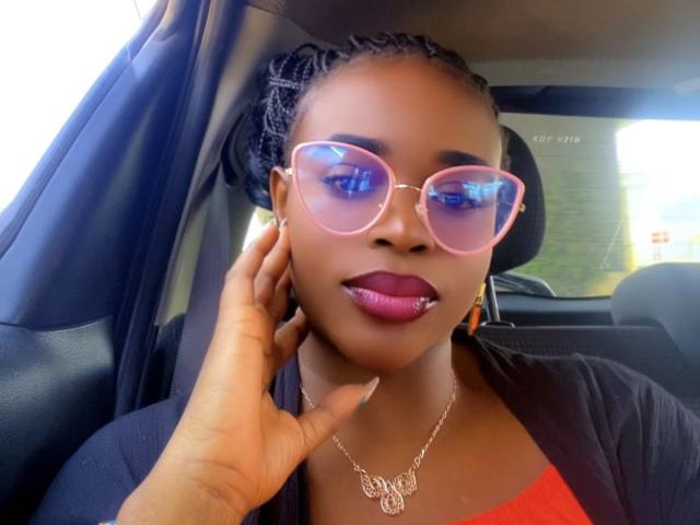 HeadLuna with dark skin, braided hair, pink reflective sunglasses, pink lipstick, silver necklace, hand touching cheek, black shirt, orange top, looking at camera, car interior.