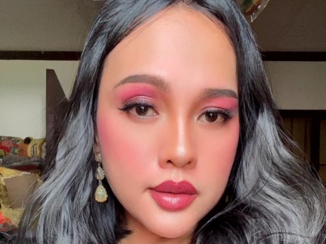 Close-up of TsPrettyNaughty with medium brown skin, black wavy hair, and pink eyeshadow. She has thick eyebrows, long eyelashes, and dark pink lipstick. Her earrings are gold with small green stones. The background is blurred, focusing on her face. The lighting is natural, highlighting her smooth skin and makeup. Her expression is neutral, with a slight smile. The image is sharp, with a slight warm tone.