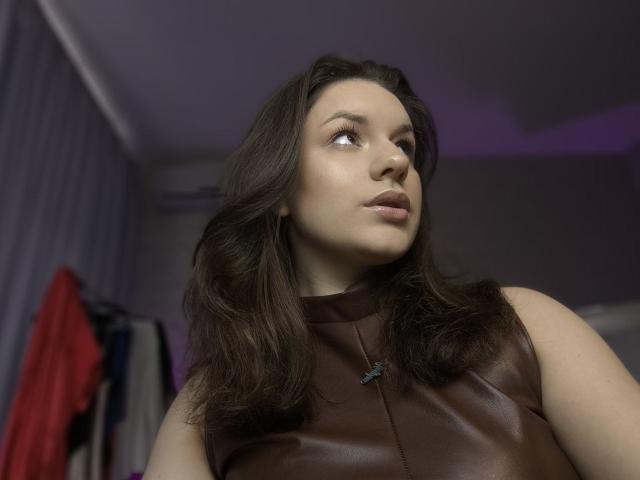 Dreia with medium brown wavy hair, brown leather sleeveless top, looking to the side, soft pink lipstick, slight smile, white skin, subtle makeup, small black necklace, indoors.