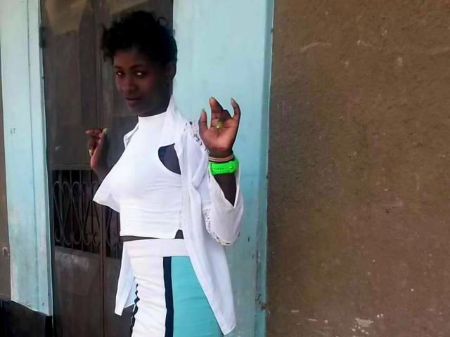 Amaja-hot with dark skin, short curly hair, wearing a white high-neck top with black cutouts and a green bracelet, standing near a door, holding a cigarette.