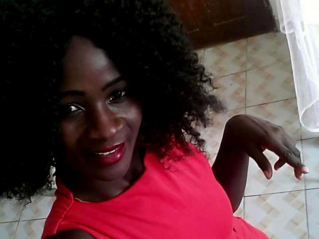 Amaja-hot with dark skin, curly black hair, red lipstick, and red dress, smiling at the camera, right hand raised, showing a finger. Boobs not visible.