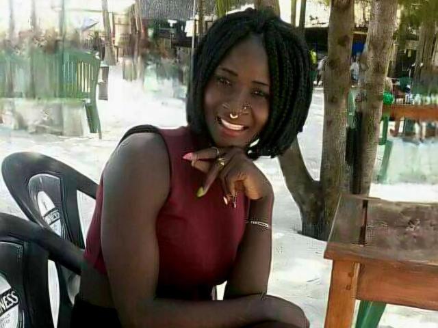 Amaja-hot with dark skin, short braids, nose ring, and smile, wearing a red sleeveless top, pointing at camera, sitting by wooden table with black chair.