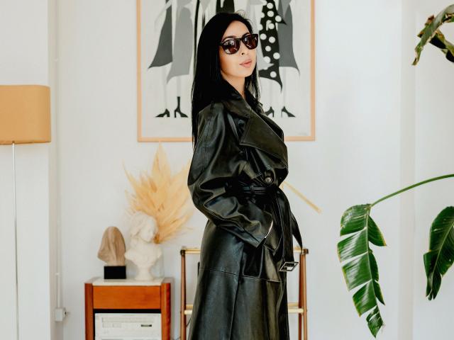 SarraRoses with black leather trench coat, hands in pockets, wearing black sunglasses, standing, looking at camera, medium skin, long black hair, small ass, pouty lips, modern room.