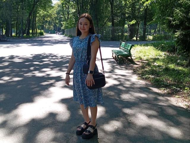 SashaDuvalle stands in a park, wearing a blue floral dress, black shoulder bag, and black platform shoes, with brown hair and a watch, under dappled sunlight.