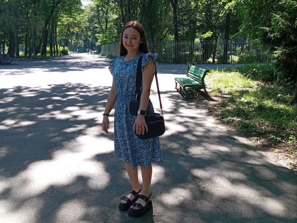 SashaDuvalle stands in a park, wearing a blue floral dress, black shoulder bag, and black platform shoes, with brown hair and a watch, under dappled sunlight.