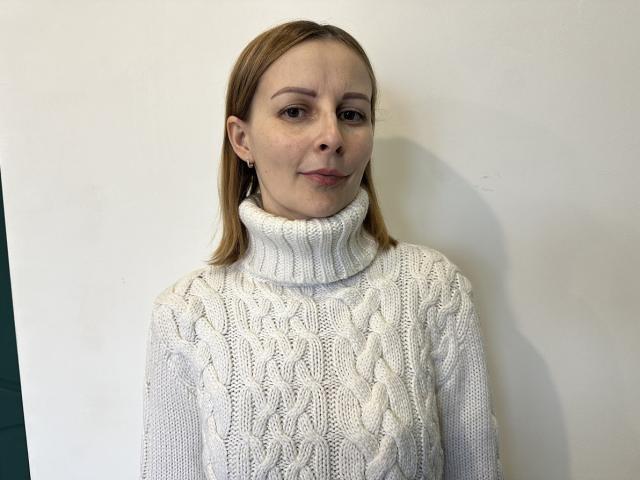 AliceMillss with pale skin, light brown hair, and subtle smile, wearing a thick white cable-knit turtleneck sweater, standing against a plain white wall.