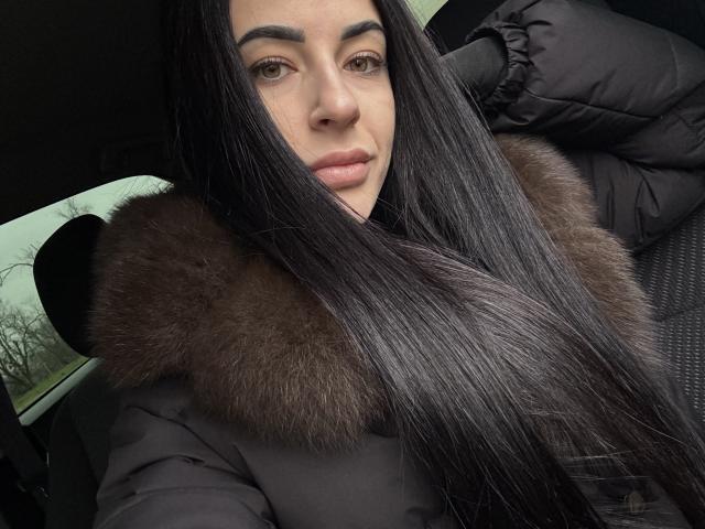 QueenViki with long black hair, brown fur-lined jacket, and pouty lips, sitting in a car, looking at the camera with a slightly confident expression.