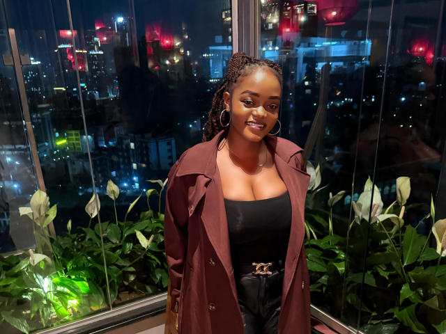 DelightfulBooby with dark skin, curly braids, hoop earrings, and a red coat over black top and pants, smiling at night in a high-rise with city lights and plants.