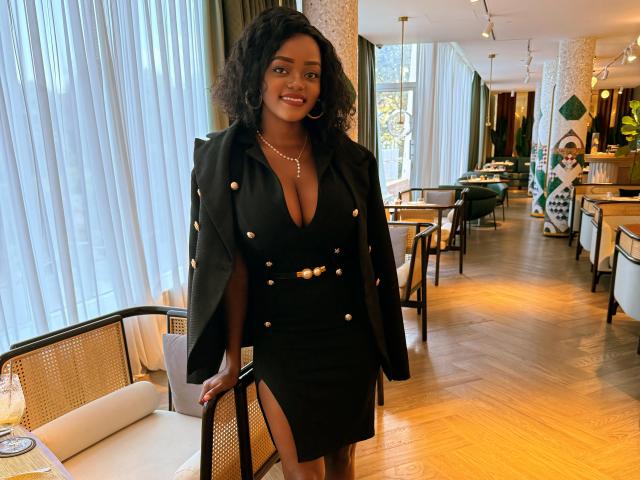 DelightfulBooby with dark curly hair, wearing a black dress with gold buttons and a matching jacket, standing in a restaurant, smiling, showing her tits in a deep V-neck.
