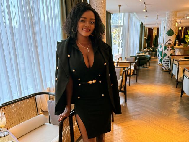 DelightfulBooby with curly black hair, wearing a black dress with gold buttons and a matching jacket, standing in a restaurant, smiling, showing a cleavage, wearing a necklace.