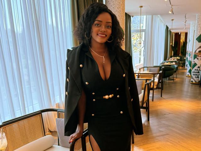 DelightfulBooby with dark curly hair, wearing a black dress with gold buttons and a deep cleavage, standing in a room with wooden floors and large windows.