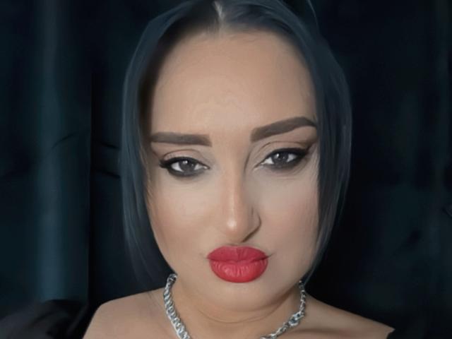 GoddessXDeborah has shiny black hair, dark eyebrows, smoky eye makeup, and bright red lips. She's wearing a silver necklace and has a serious expression.