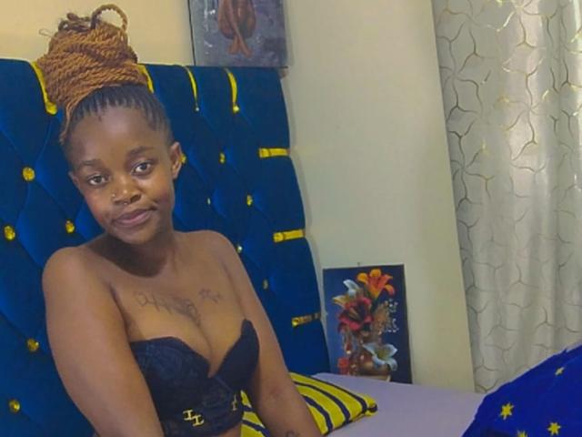 SpicyPrettyx with dark skin, small tattoos above her tits, in a black lace strapless top, brown braided bun, blue velvet headboard, and yellow-patterned pillow, smiling slightly.