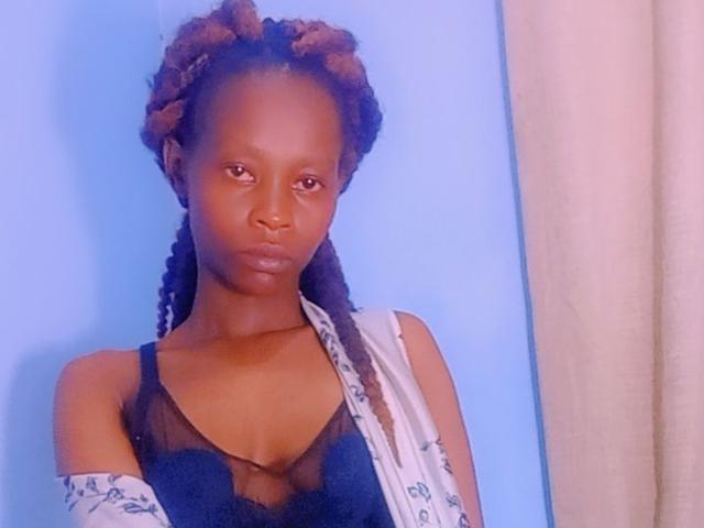 Photograph of PelgyCarter. She has dark brown skin and curly, twisted hair with a reddish tint. She wears a black lace bralette with a sheer fabric and a white scarf with blue patterns. Her expression is neutral, with slightly parted lips. She's in a blue background with a beige curtain on the right. The image is slightly blurry. She has a slender build and small breasts. Her gaze is directed at the camera.